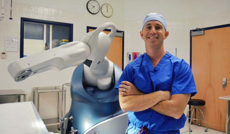 Dr. Scott Goldsmith posing in front of the robot used to assist in joint replacement procedures Post